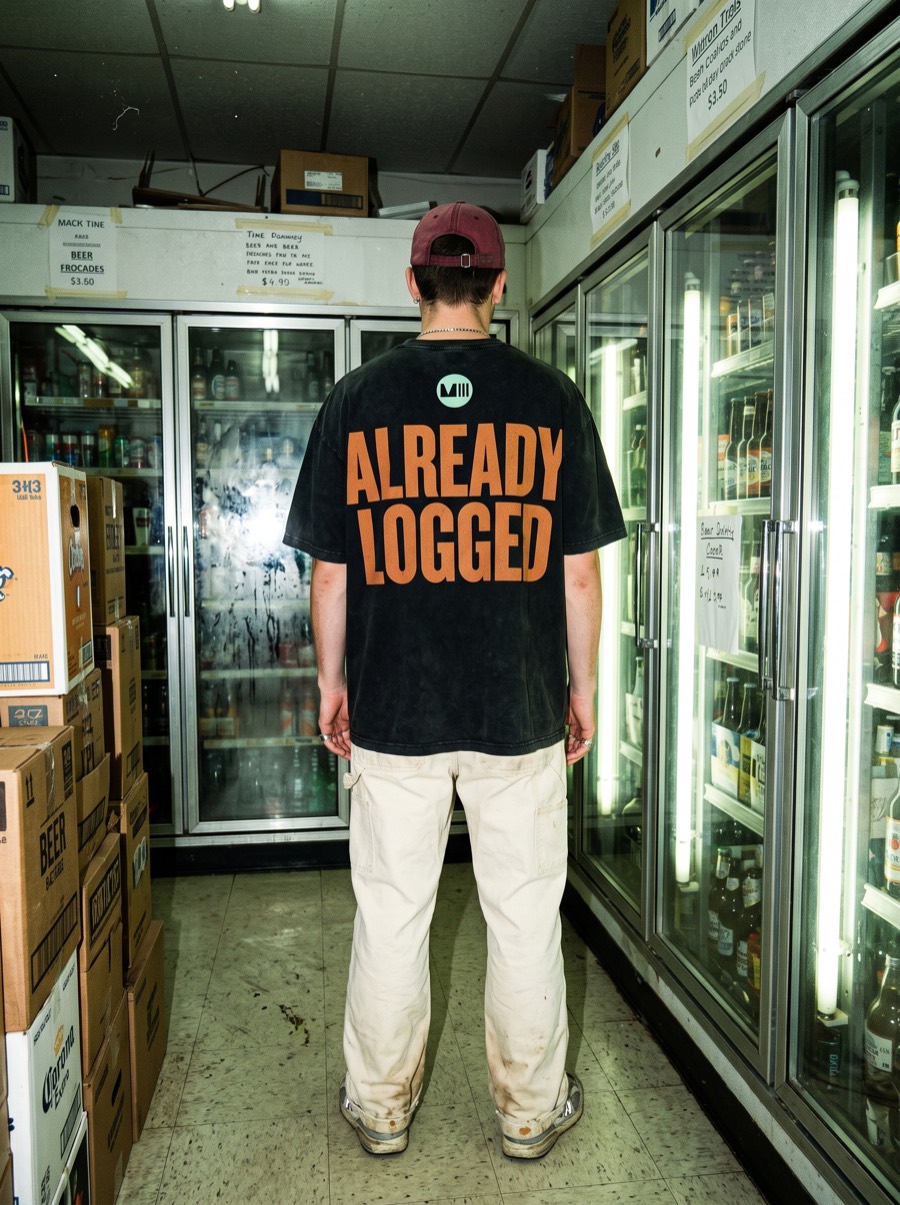 A man in an ALREADY LOGGED T-shirt standing at the cooler doors of a corner shop, his food tracked before he decides