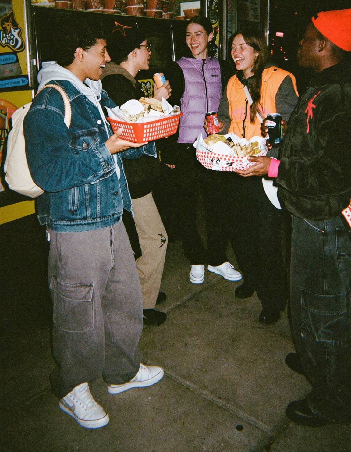 Five friends laughing together outside a food truck at night, holding baskets of food and cans